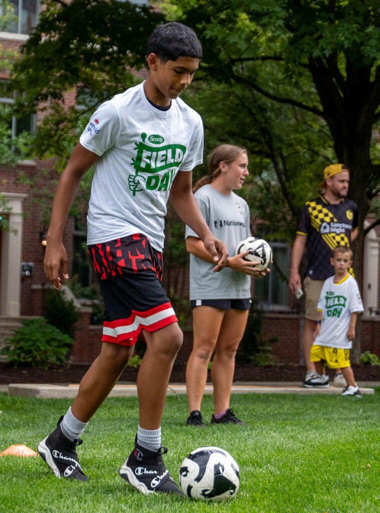 Photo of a kid kicking a soccer ball on green grass with a few people in the background