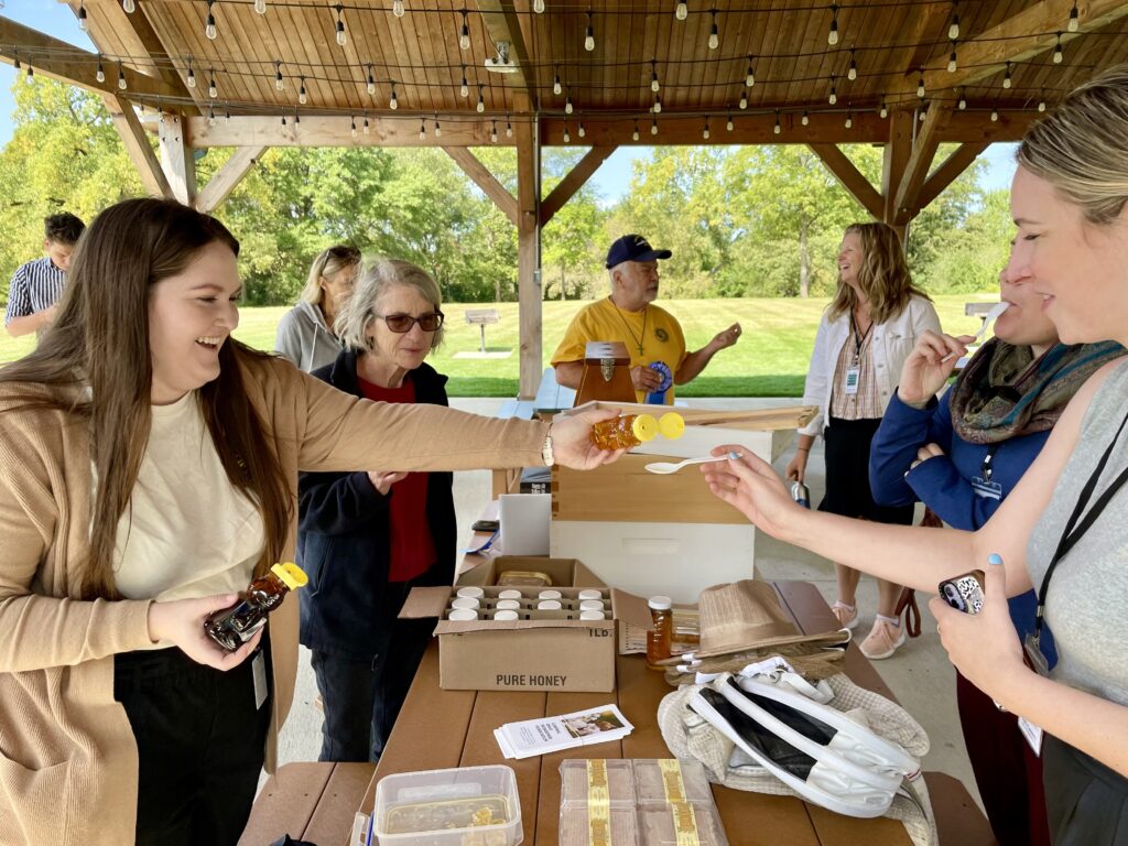 People sampling honey under a pavilion with green trees and grass behind them