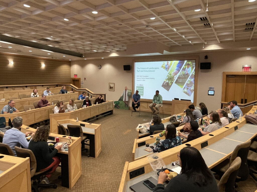 Image of a large arena meeting room with people viewing a presentation made by two people at the front of the room