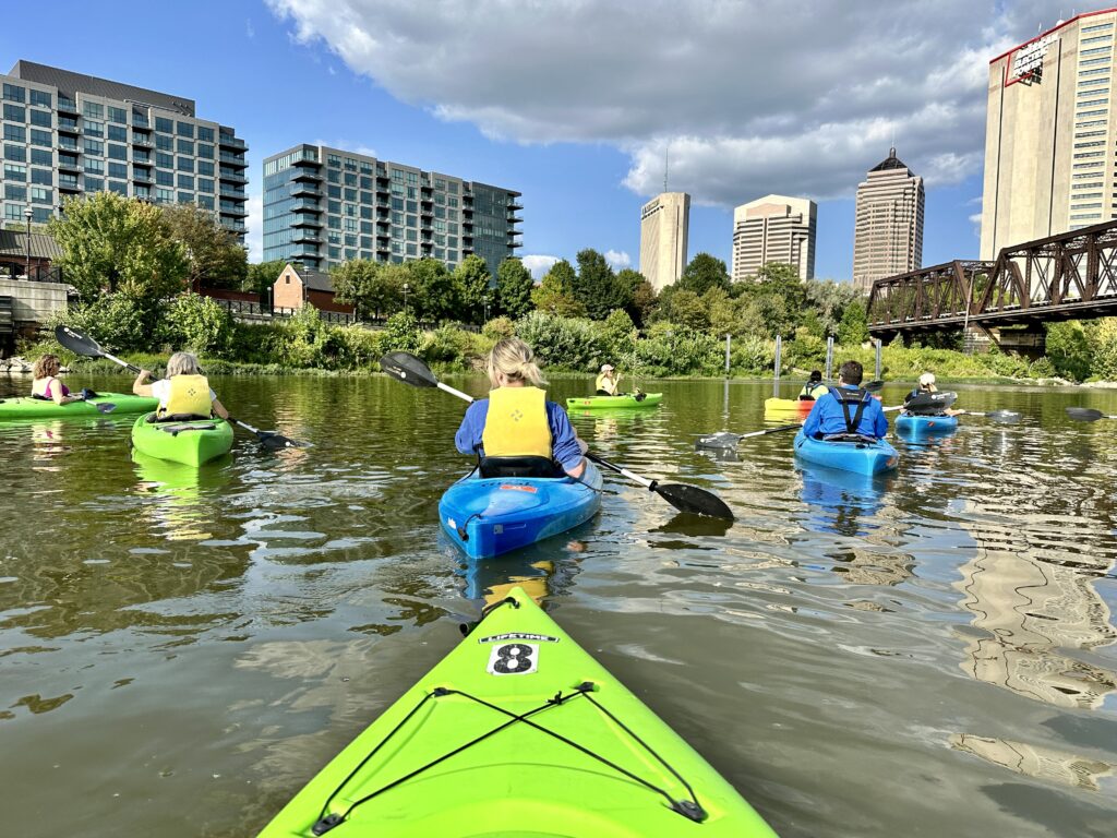 Several kayaks on a river with buildings and green plants in the background